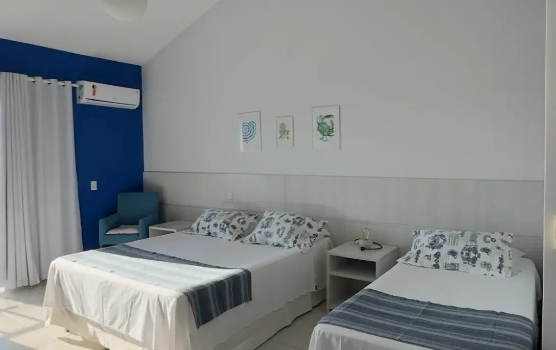 Quarto com duas camas individuais, parede azul, pendente de teto, três quadros decorativos, ar condicionado e cortinas brancas