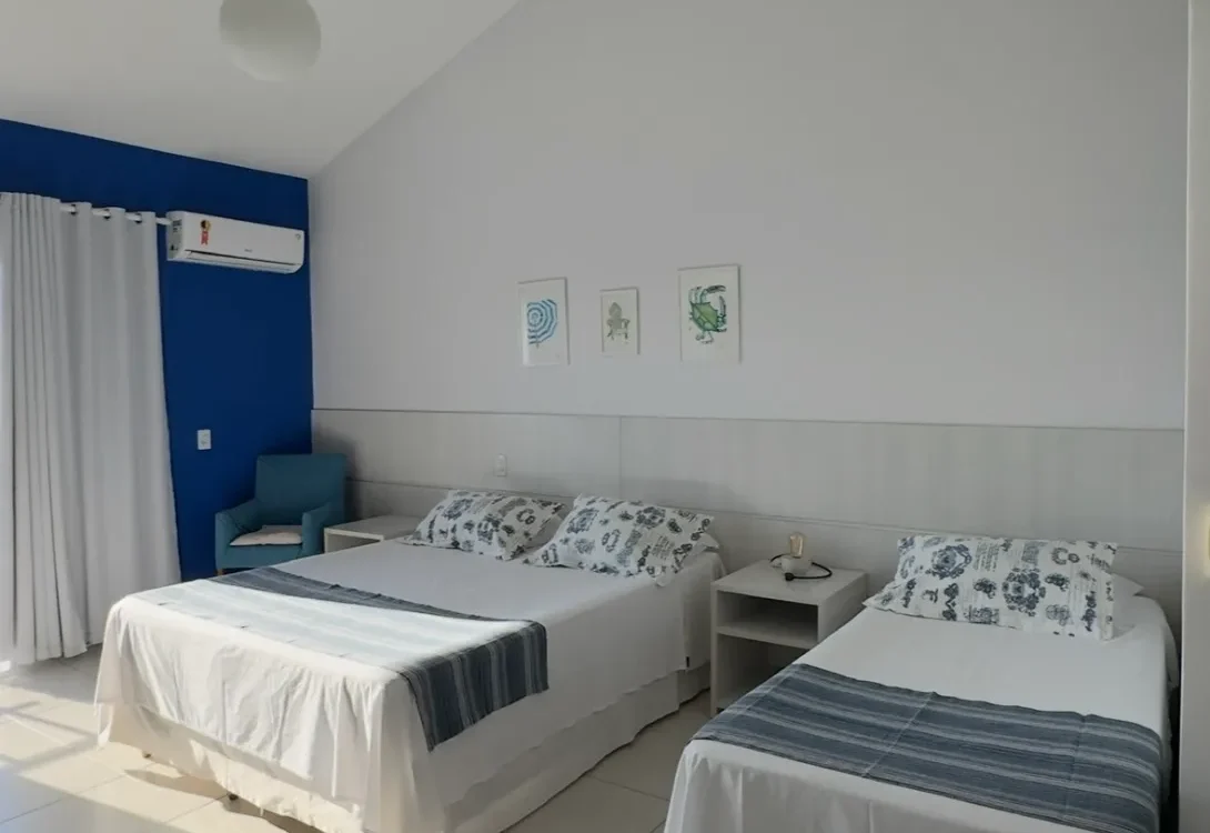 Quarto com duas camas individuais, parede azul, pendente de teto, três quadros decorativos, ar condicionado e cortinas brancas